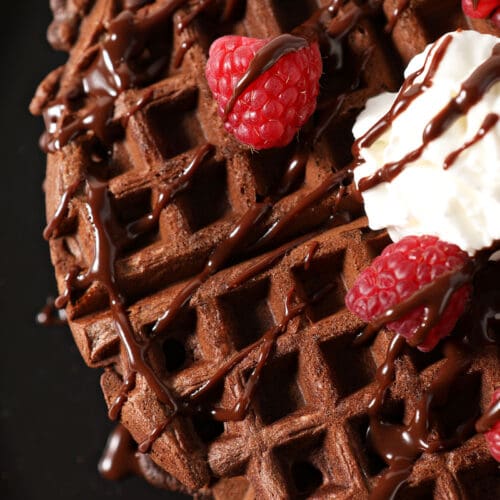 Overhead of a chocolate waffle drizzled with chocolate sauce, strawberries and whipped cream on a black plate