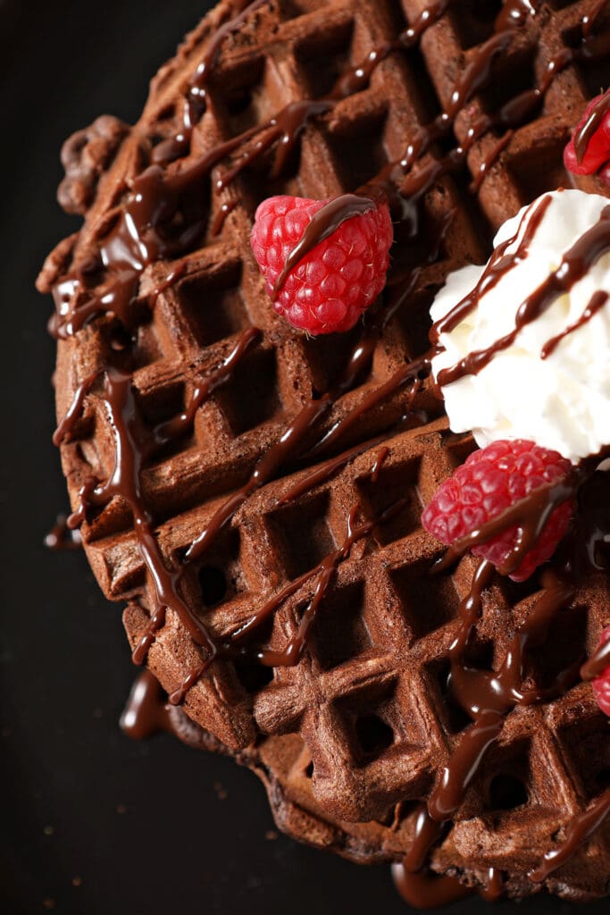 Overhead of a chocolate waffle drizzled with chocolate sauce, strawberries and whipped cream on a black plate