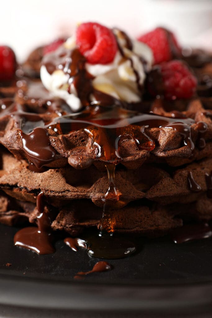 Close up of maple syrup dripping down a stack of chocolate waffles topped with whipped cream, raspberries and chocolate sauce