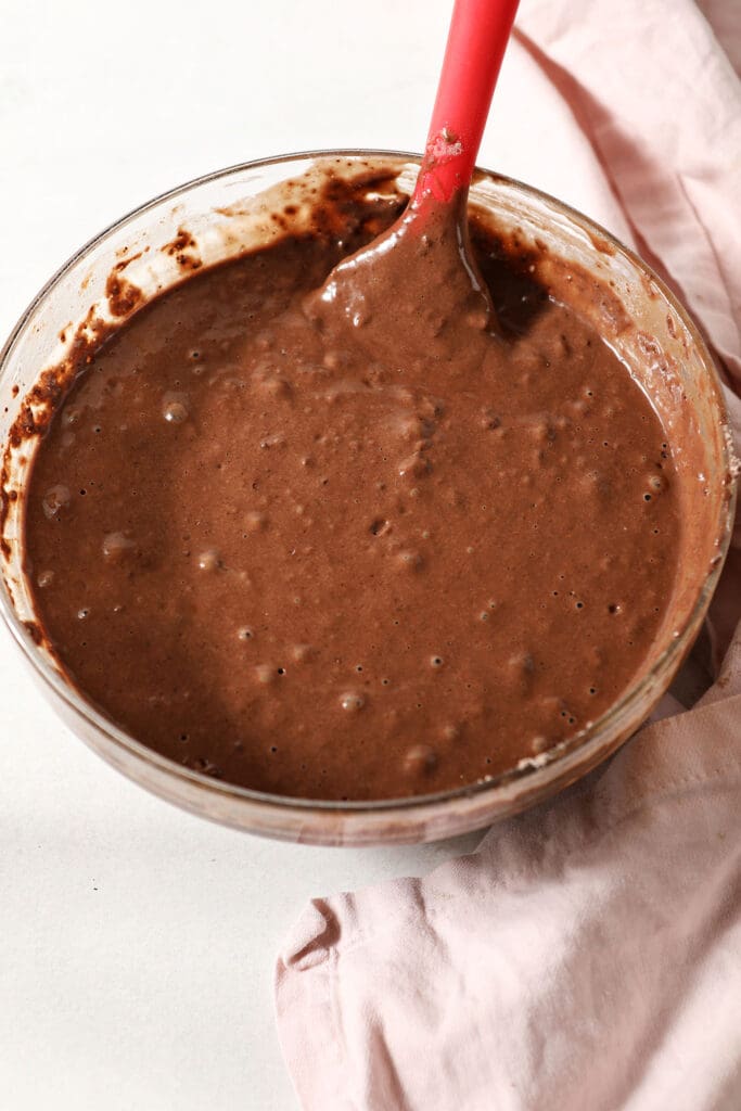 Chocolate waffle batter in a bowl with a red spatula on marble with a pink linen next to it