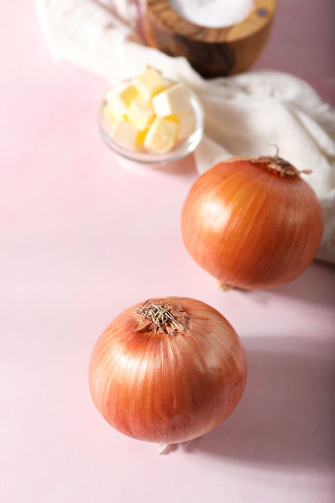 Two onions, a bowl of butter and salt on a pink surface with a white linen