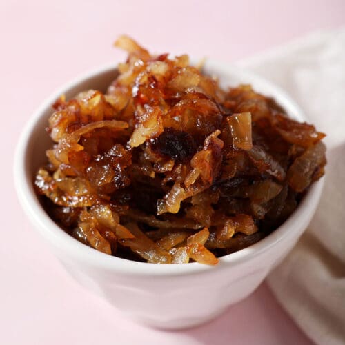 A bowl of caramelized onions on a pink surface next to a white linen