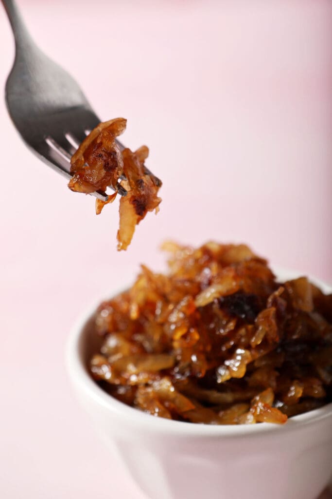 A fork holds a bite of caramelized onions above a bowl of onions on a pink surface