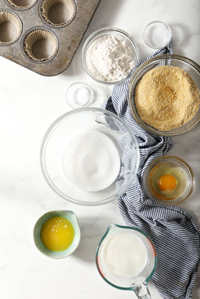 Ingredients to make cornbread muffins in bowls with a blue and white striped towel on marble