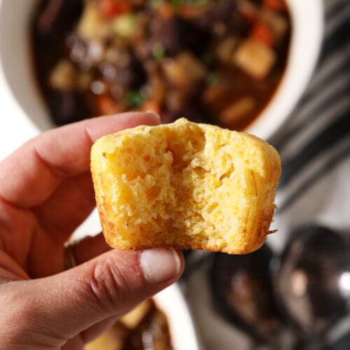 A hand holds a bitten-into cornbread muffin over bowls of beef stew