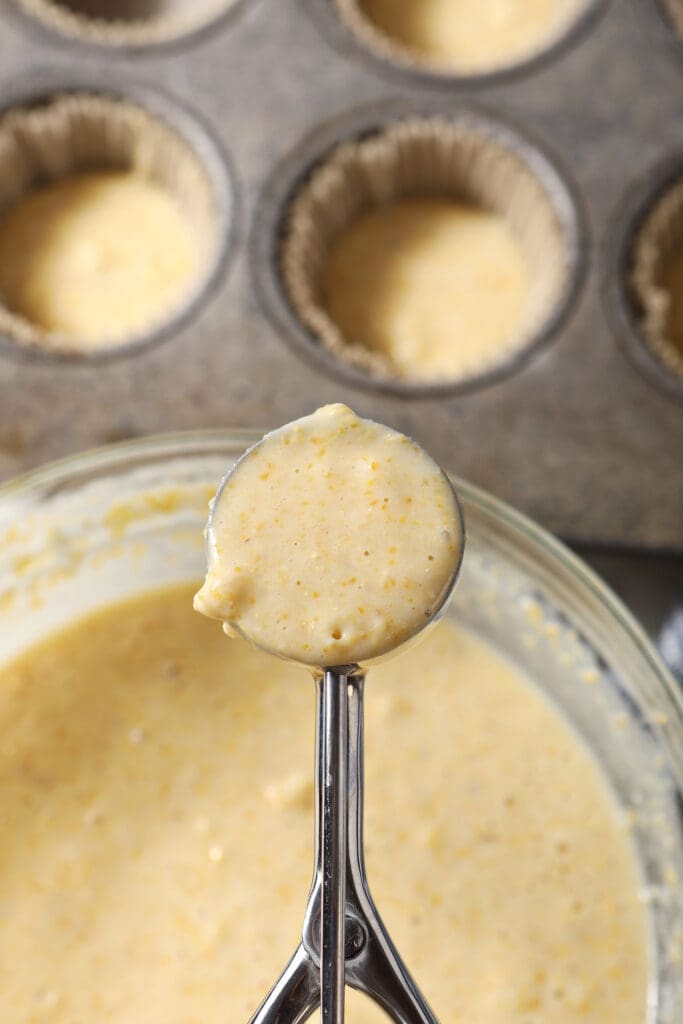 A cookie scoop holds muffin batter above a bowl with more batter and a muffin tin