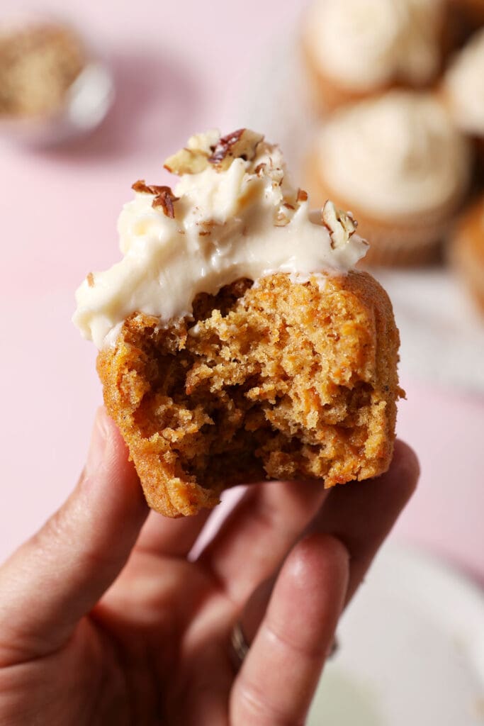 A hand holds a bitten-into Carrot Cupcake with cream cheese frosting