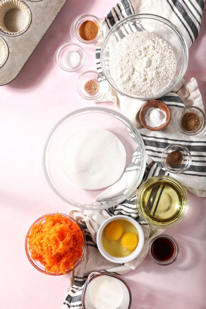 Bowls of ingredients to make carrot cupcakes on a pink surface with a striped black and white linen