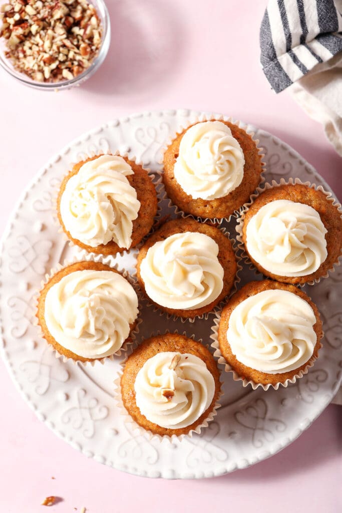 Frosted carrot cupcakes on a white plate on a pink surface with a bowl of chopped nuts next to them