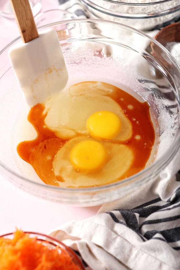 Eggs and vanilla are added to wet ingredients in a bowl on a pink surface