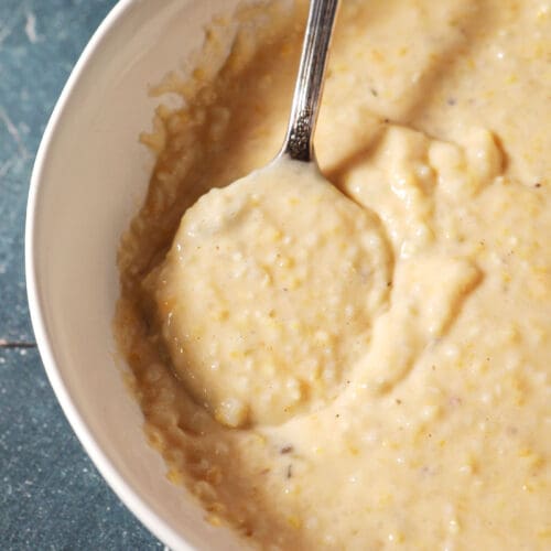 a spoon sits in a white bowl of cheese grits on a dark green surface