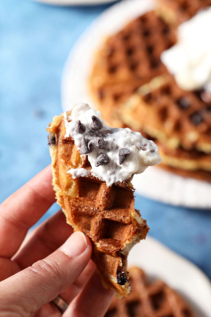 a hand holds a bitten-into waffle dipped in whipped cream and covered in chocolate chips over a plate holding another waffle on a blue surface