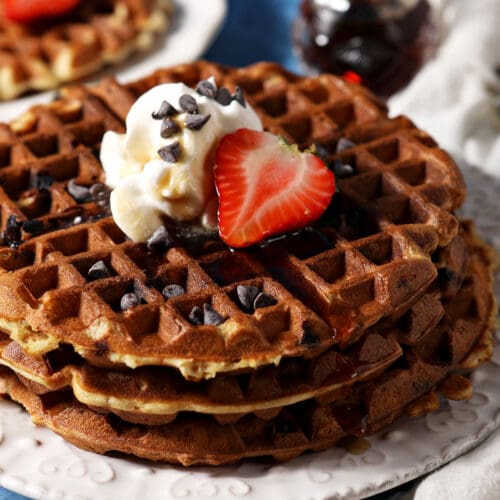 A stack of three chocolate chip waffles topped with whipped cream and strawberries on a white plate