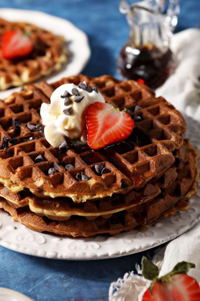 A stack of three chocolate chip waffles topped with whipped cream and strawberries on a white plate