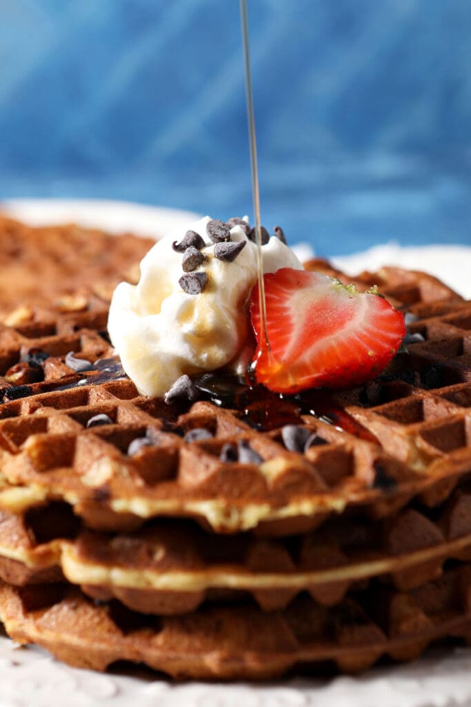 syrup pours on top of a stack of chocolate chip waffles topped with a halved strawberry and whipped cream