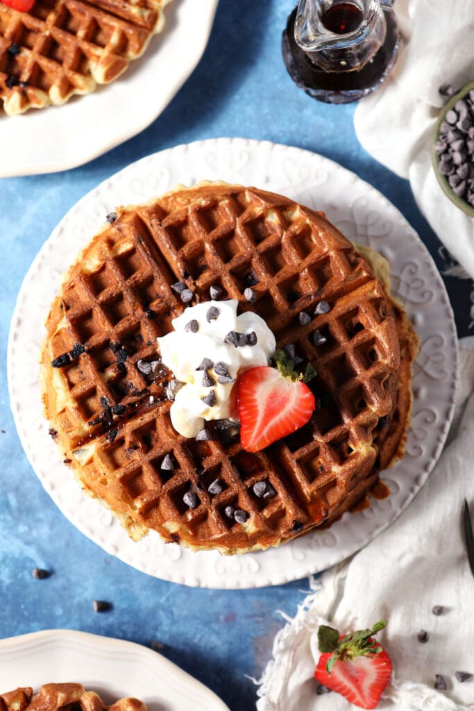 overhead of a waffle on a white plate topped with whipped cream, chocolate chips and a halved strawberry on a blue surface