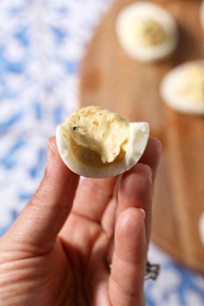 A hand holds a bitten-into deviled egg over a board of more eggs on a blue and white tiled surface