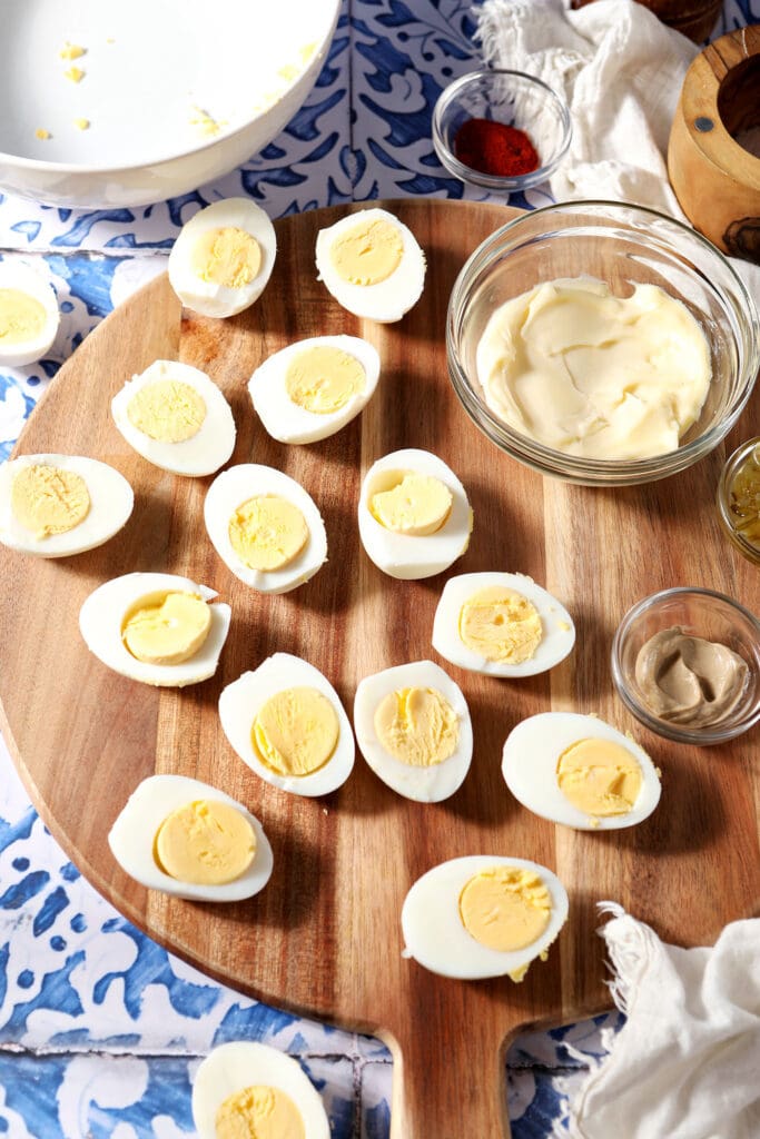 Halved hard boiled eggs with other deviled egg ingredients in bowls on a wooden platter on a blue and white tiled surface