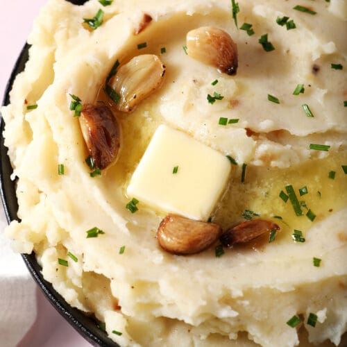 close up overhead of a bowl of roasted garlic mashed potatoes topped with melted butter and roasted garlic cloves