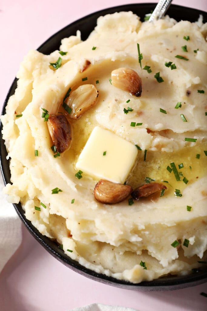 close up overhead of a bowl of roasted garlic mashed potatoes topped with melted butter and roasted garlic cloves