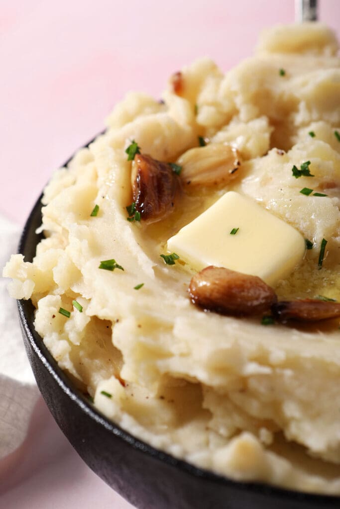 close up of roasted garlic mashed potatoes topped with melted butter and roasted garlic in a bowl on a pink surface