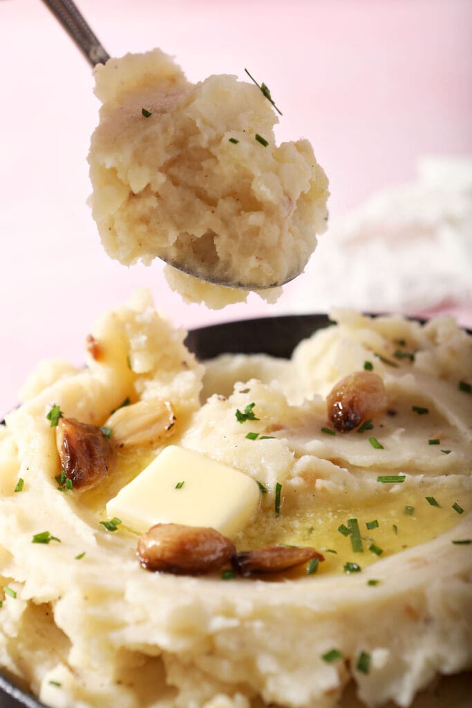 a spoon lifts mashed potatoes out of a bowl on a pink surface surrounded by white linens