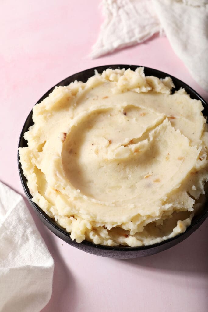 roasted garlic mashed potatoes in a black bowl on a pink surface surrounded by white linens