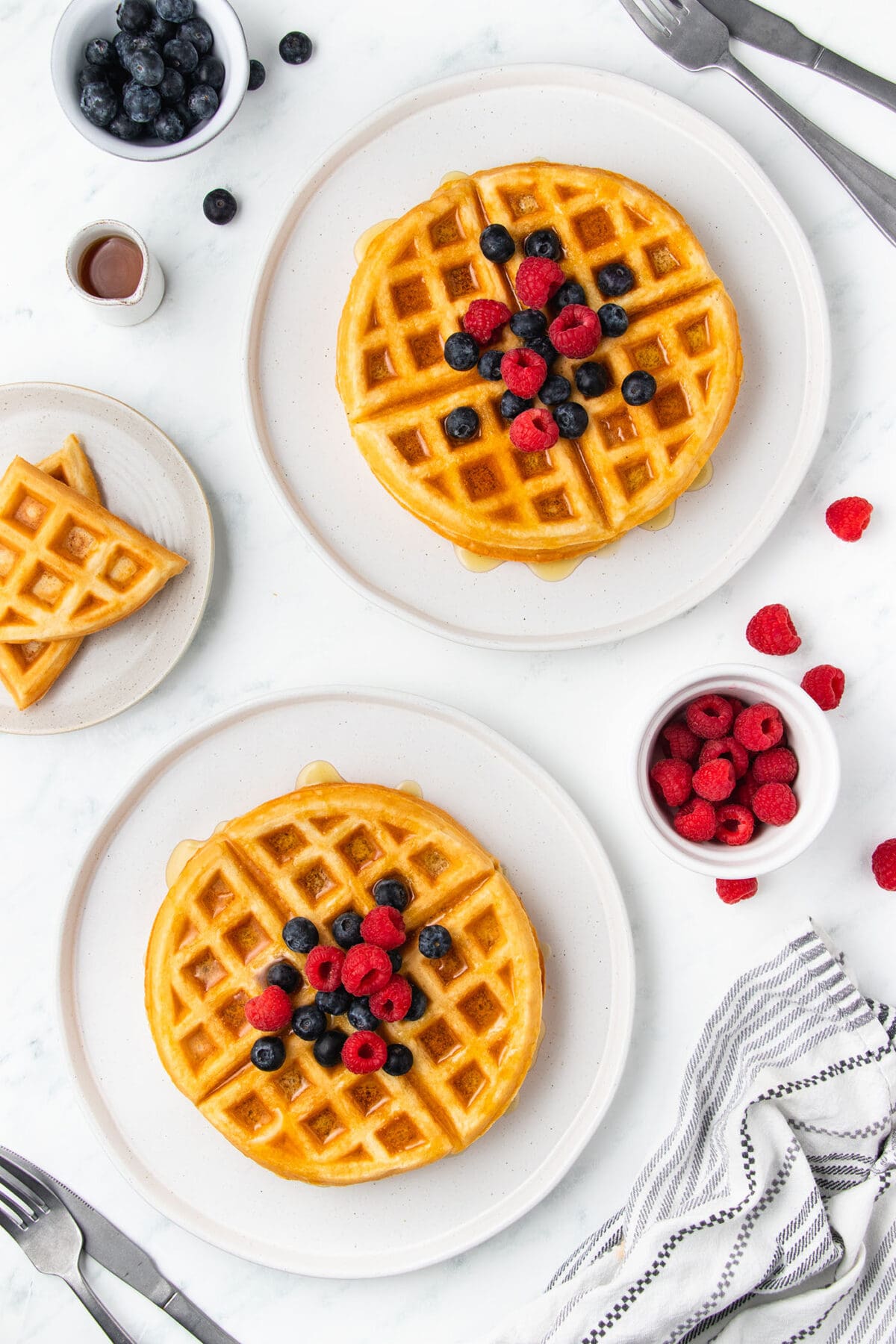 overhead image of two buttermilk waffles topped with berries and syrup on white plates on a white surface