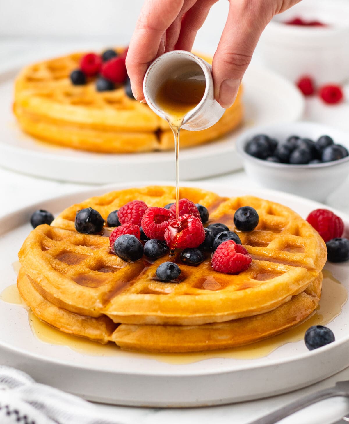 maple syrup pours on top of a waffle on a white plate topped with berries