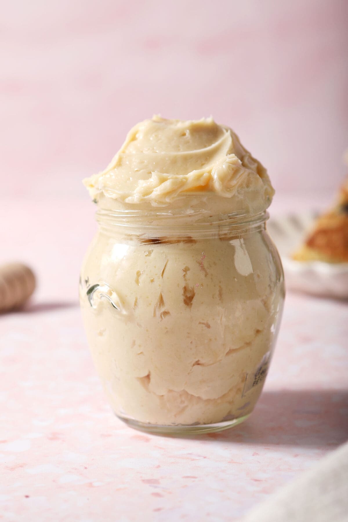 A glass jar of whipped honey butter on a pink surface in front of a plate of biscuits
