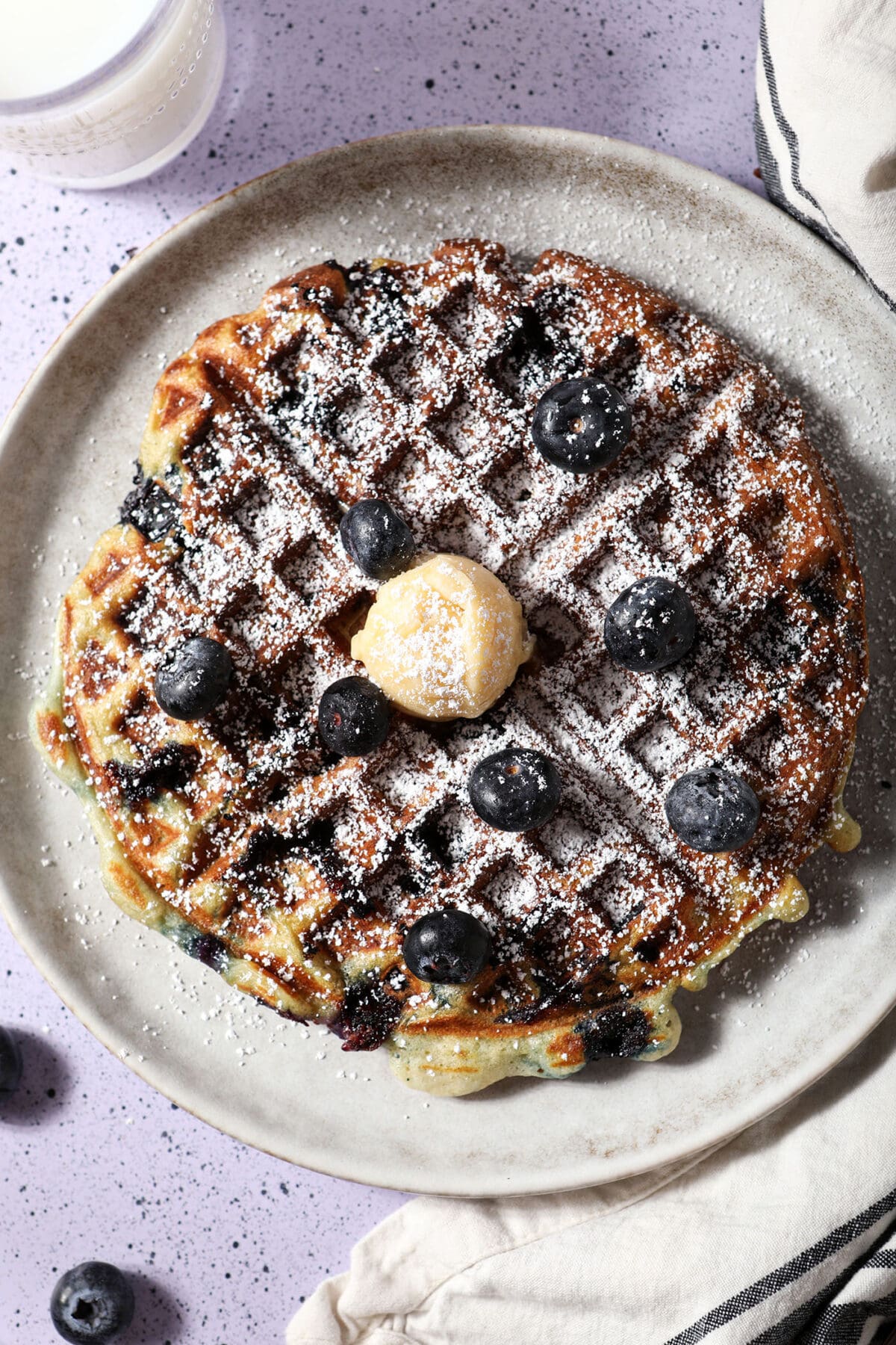 A blueberry waffle topped with powdered sugar, fresh blueberries and a dollop of honey butter on a gray plate on a purple speckled surface