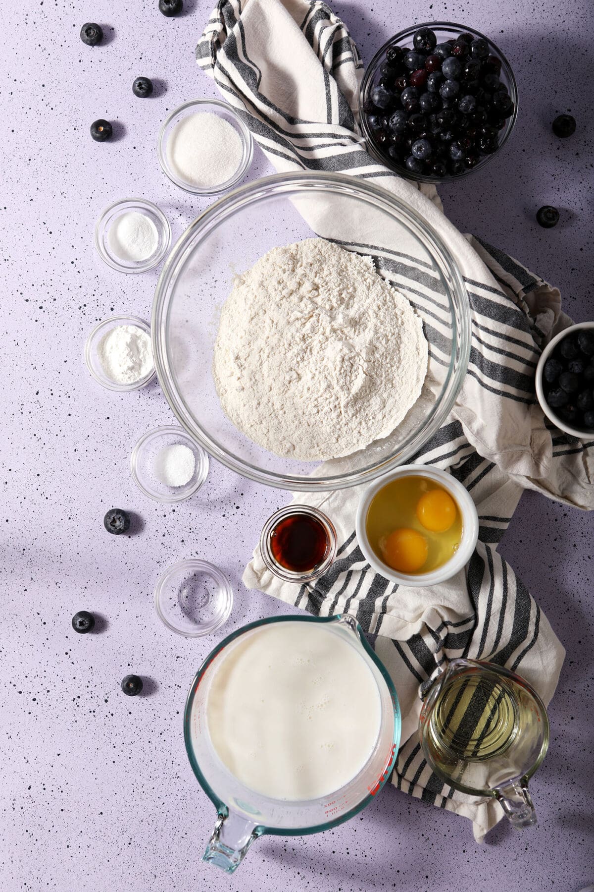Ingredients to make waffles with blueberries on a purple speckled surface with a white and black striped linen