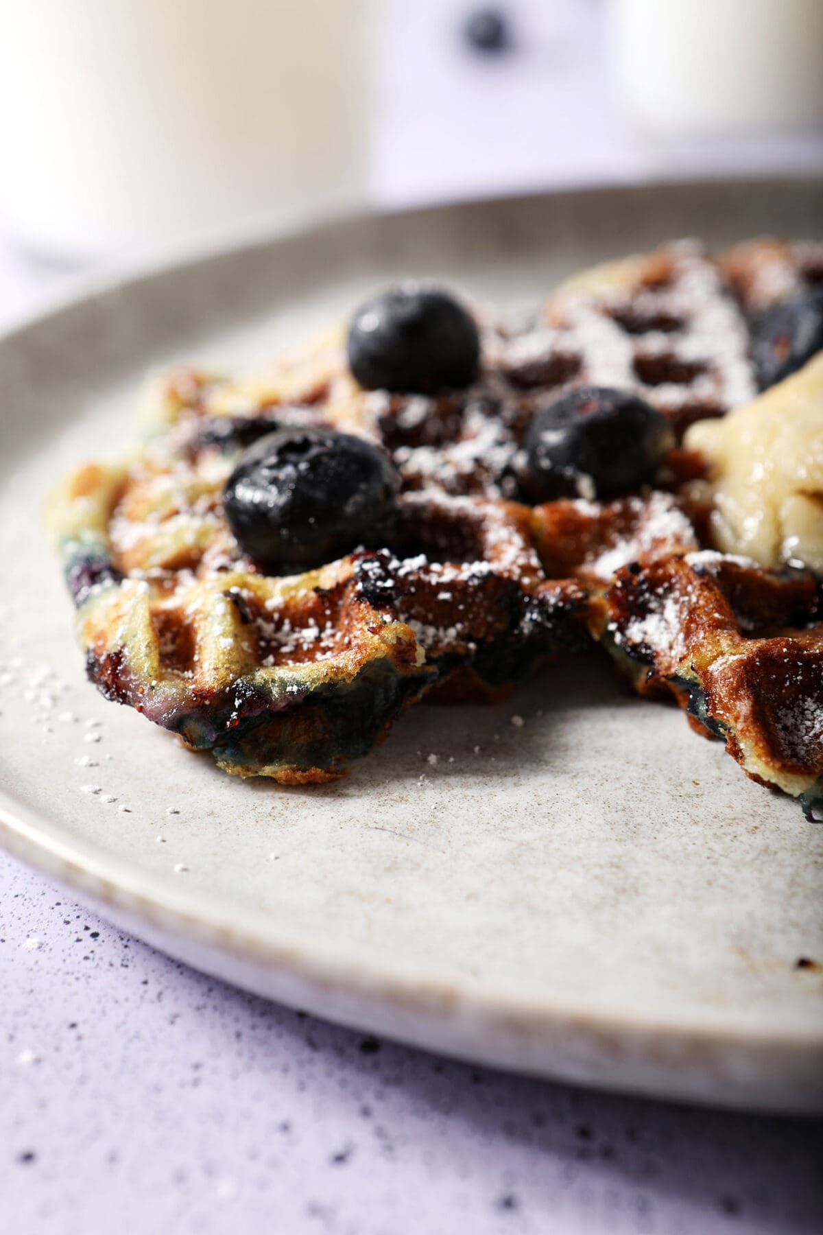 Side angle of a blueberry waffle topped with extra blueberries on a gray plate on a purple speckled surface
