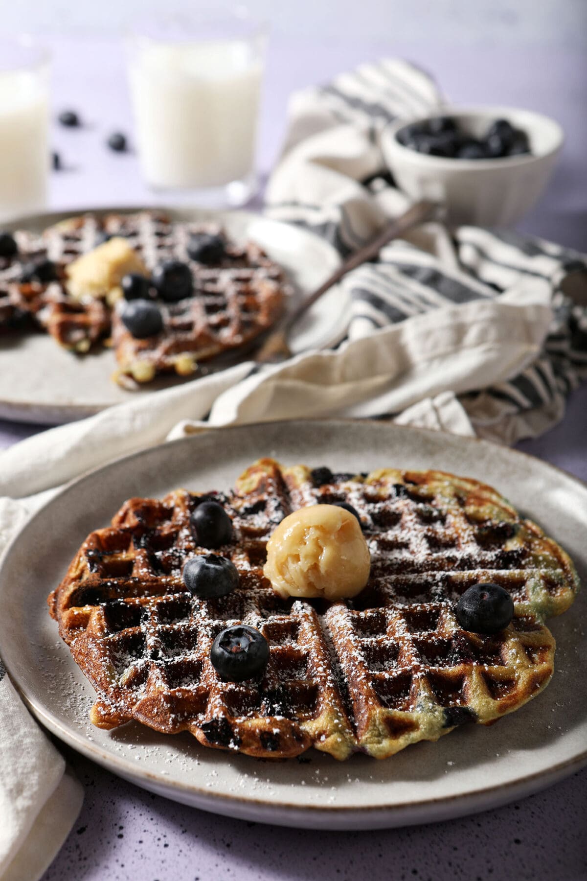 Two gray plates of blueberry waffles topped with butter and blueberries on a purple surface with cups of milk and a bowl of blueberries