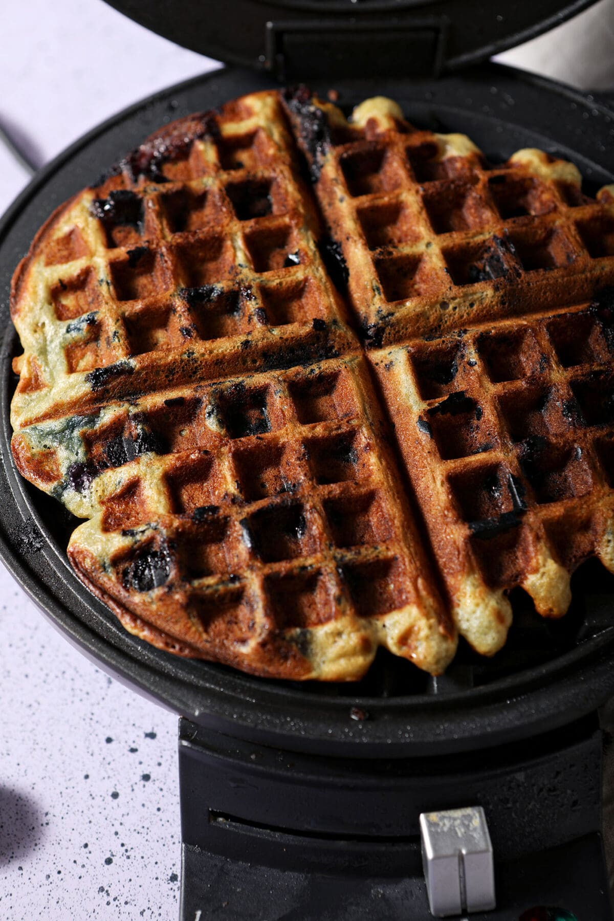 A blueberry waffle in a waffle iron on a purple speckled surface