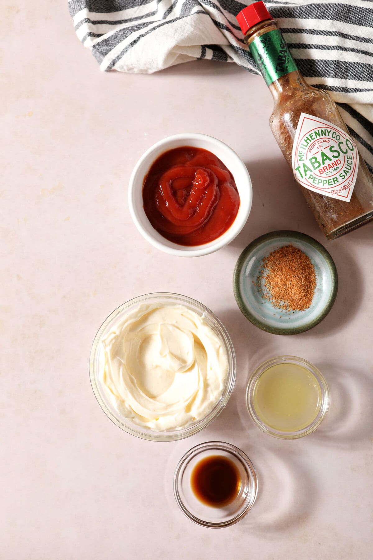 Bowls holding ingredients to make Cajun dipping sauce on a light taupe surface next to a white and black striped linen