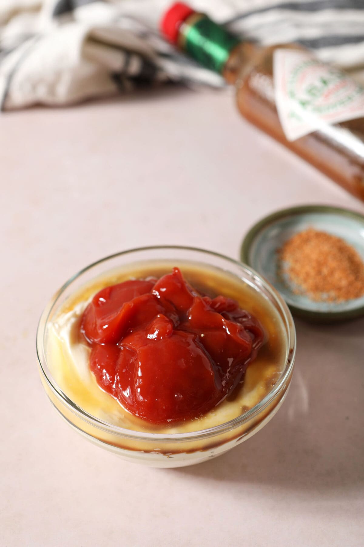 Ingredients for cajun dipping sauce partially poured into a small glass bowl on a taupe surface