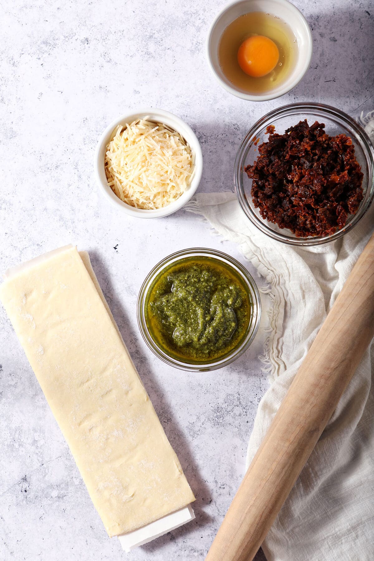 Ingredients to make pesto pinwheels with puff pastry on a grey surface in bowls