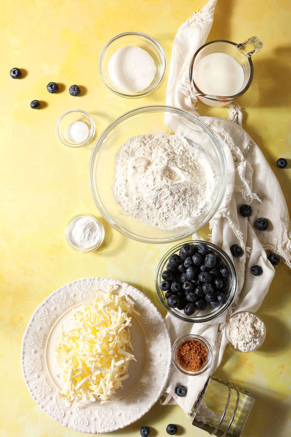 Ingredients to make buttery biscuits with blueberries in bowls on a yellow surface with a white linen