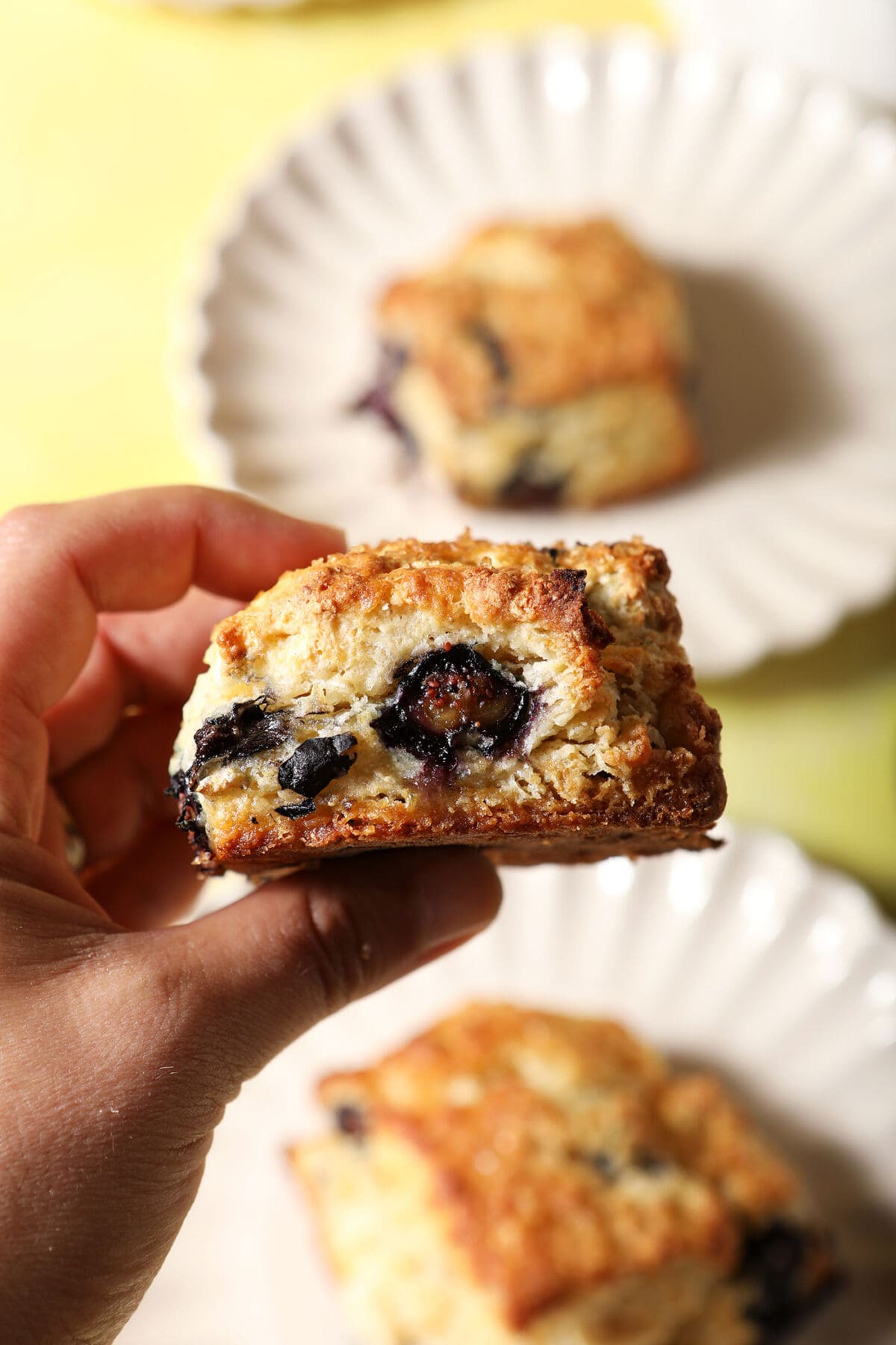 A hand holds a blueberry biscuit above two other plates of biscuits