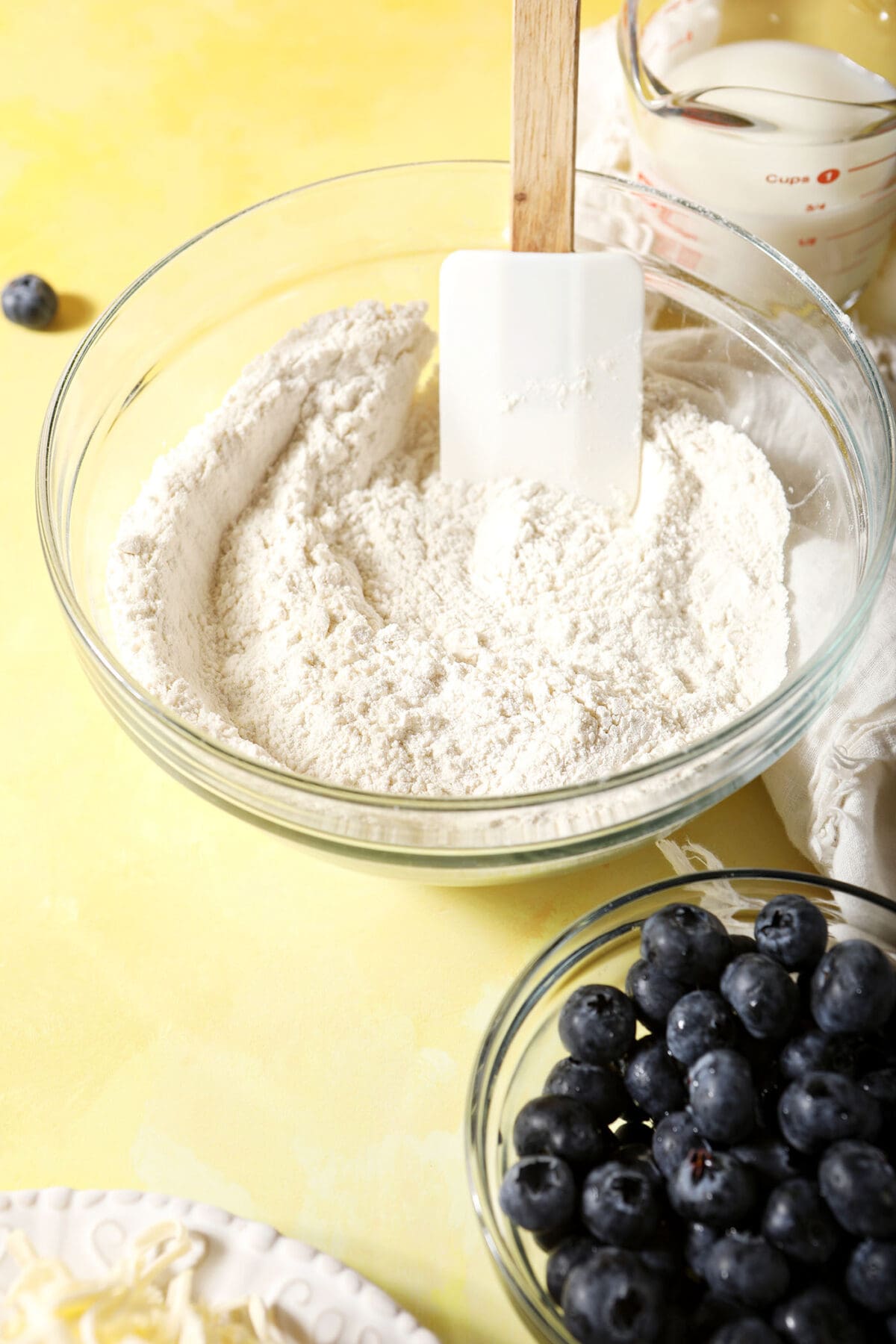 A bowl of dry ingredients with a rubber spatula in it next to a bowl of blueberries on a yellow surface