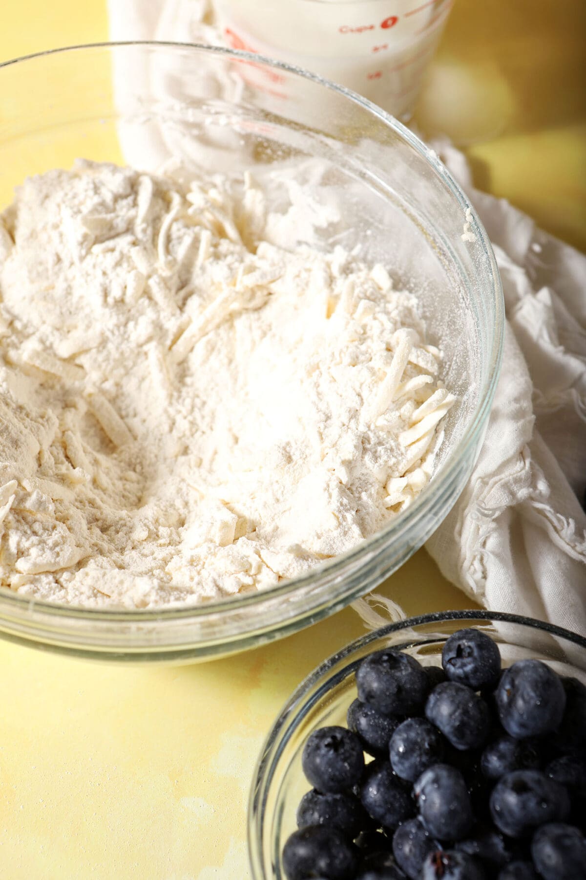 A bowl of dry ingredients with shredded butter next to a bowl of blueberries on a yellow surface