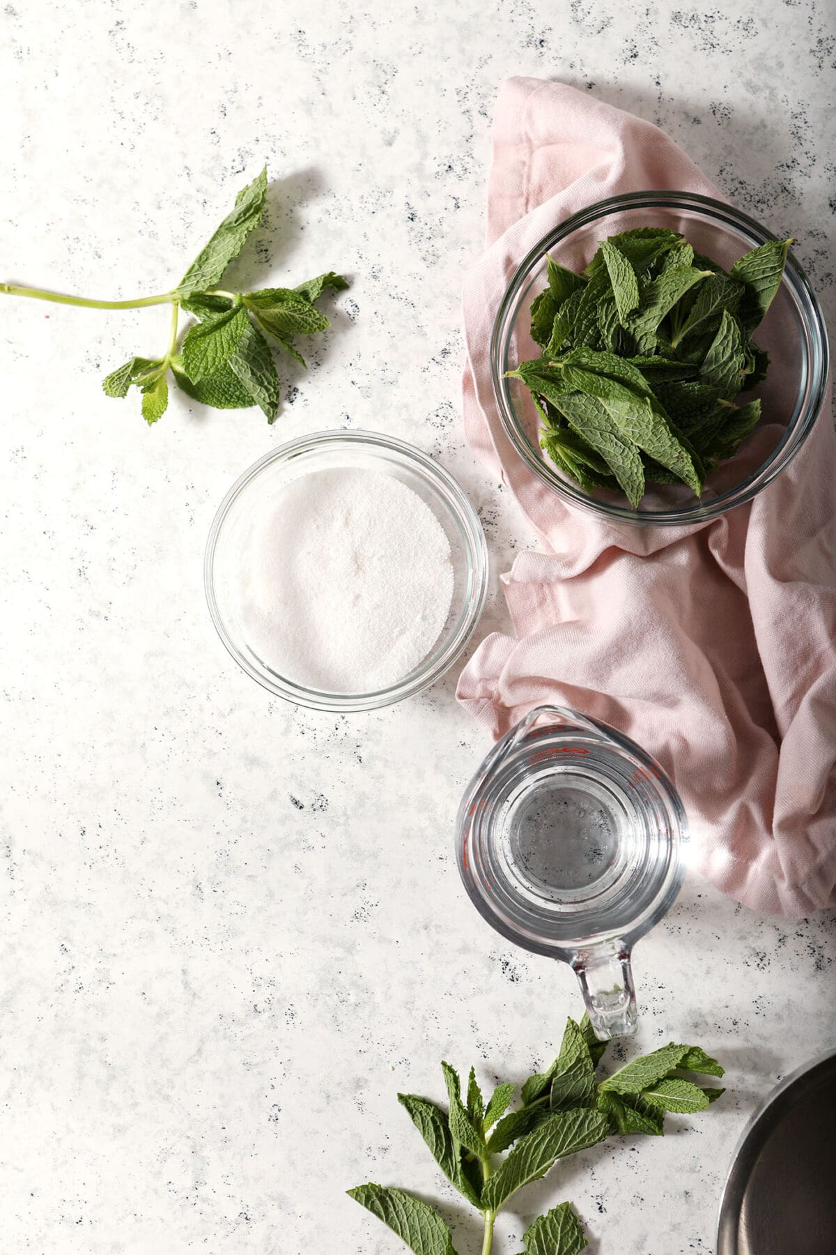 ingredients to make mint syrup in bowls on a marble surface next to a pink linen and fresh mint leaves