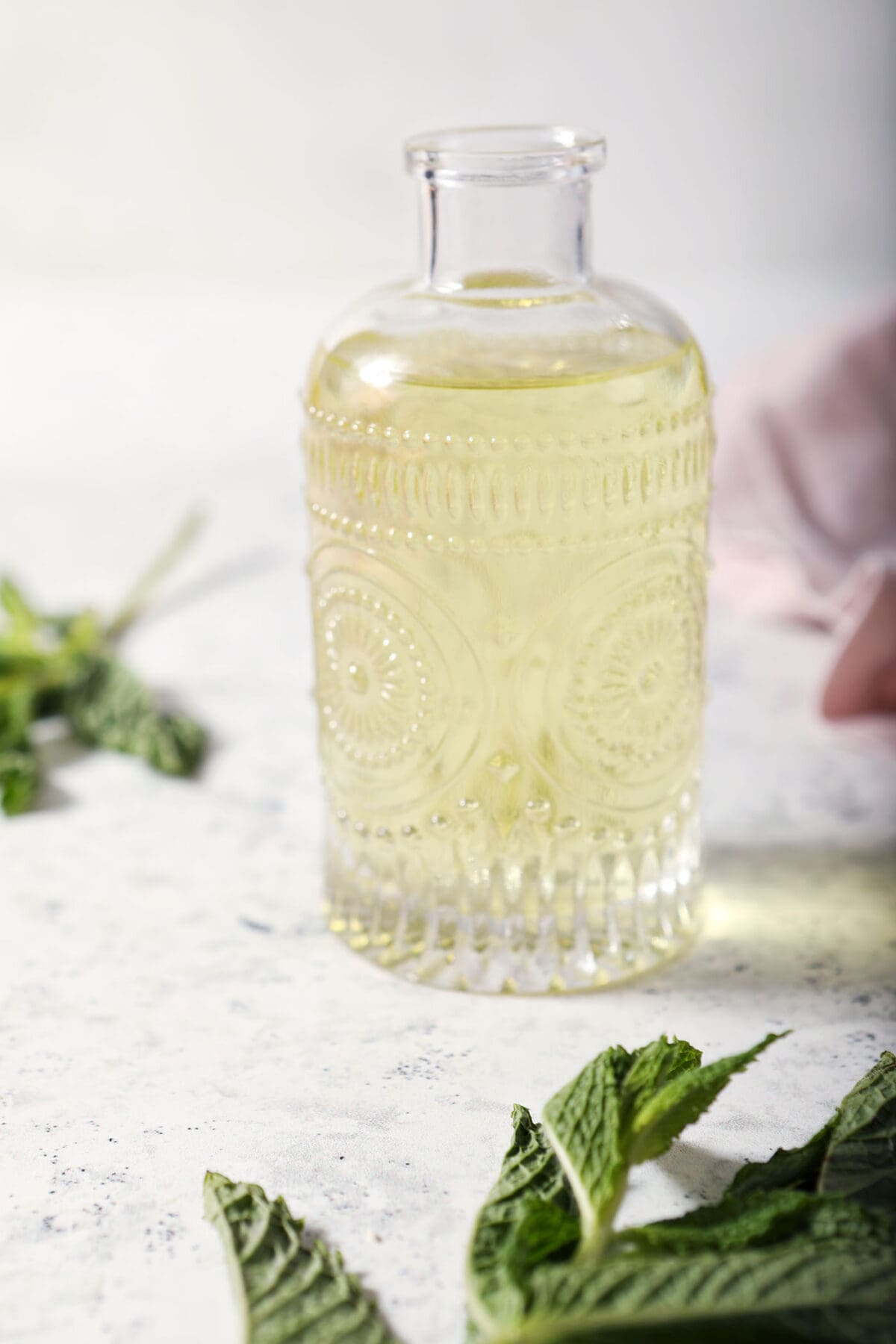 close up of a bottle of mint simple syrup sits on a marble surface surrounded by mint leaves and a pink linen