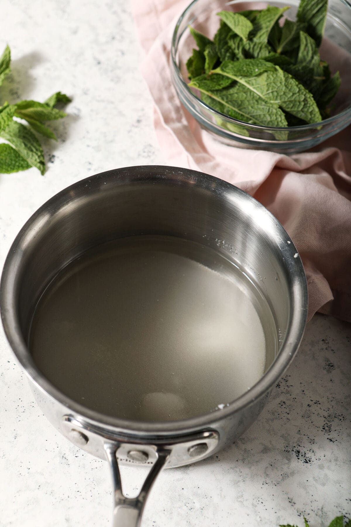 water and sugar in a small saucepan on a marble surface surrounded by mint leaves