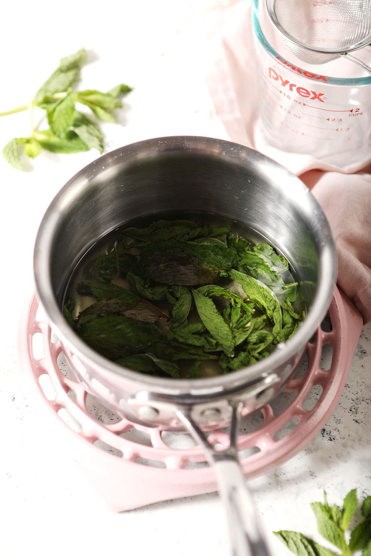 close up of mint leaves infuse in a saucepan of simple syrup on a pink trivet on stone surrounded by fresh mint leaves
