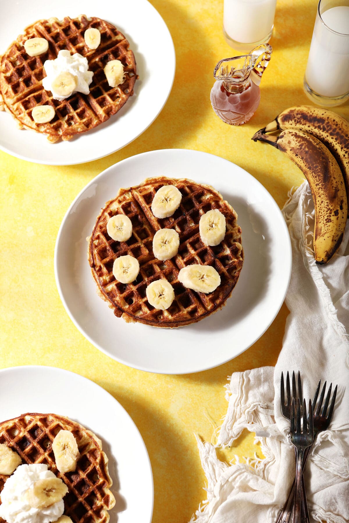 Three banana waffles on white plates on a yellow surface next to bananas, forks and a white linen