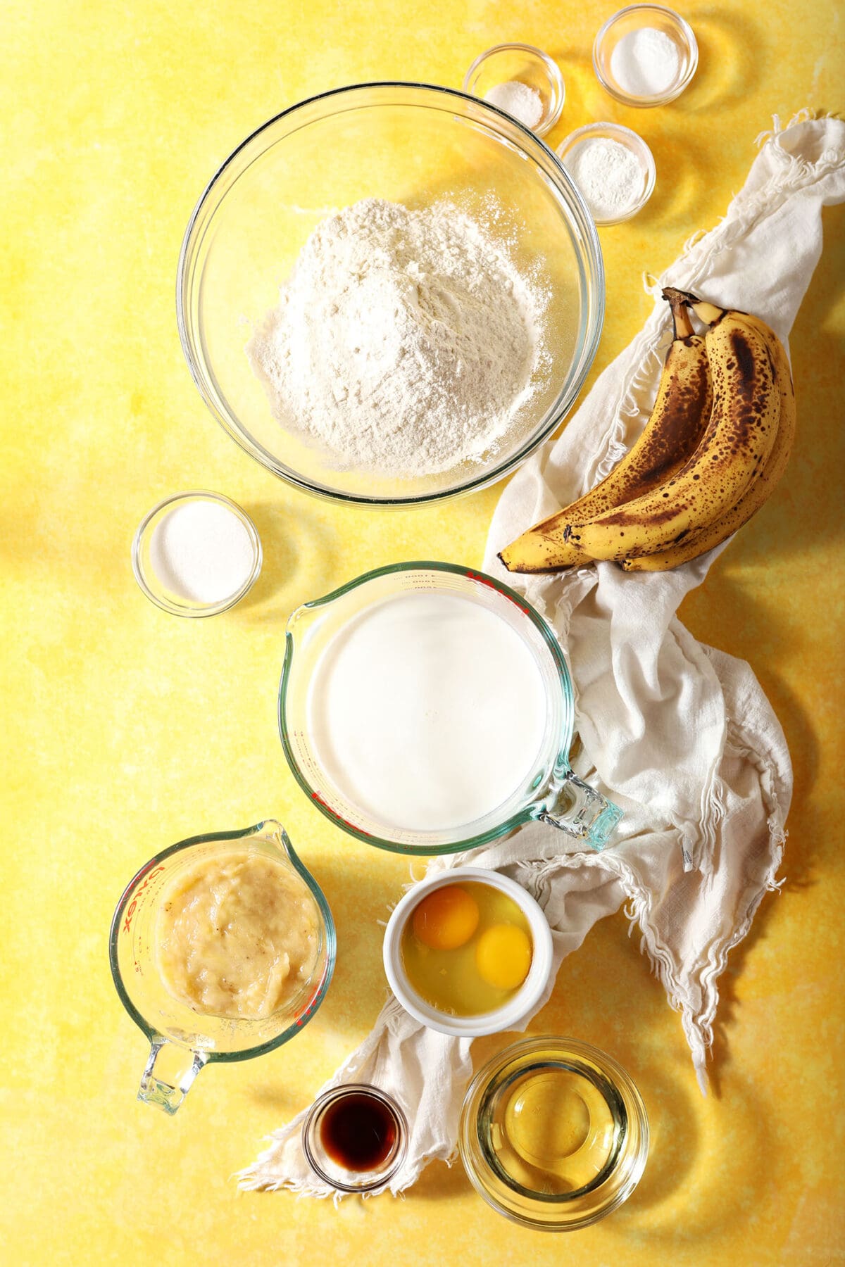Ingredients in bowls to make banana waffles on a yellow surface next to bananas and a white linen