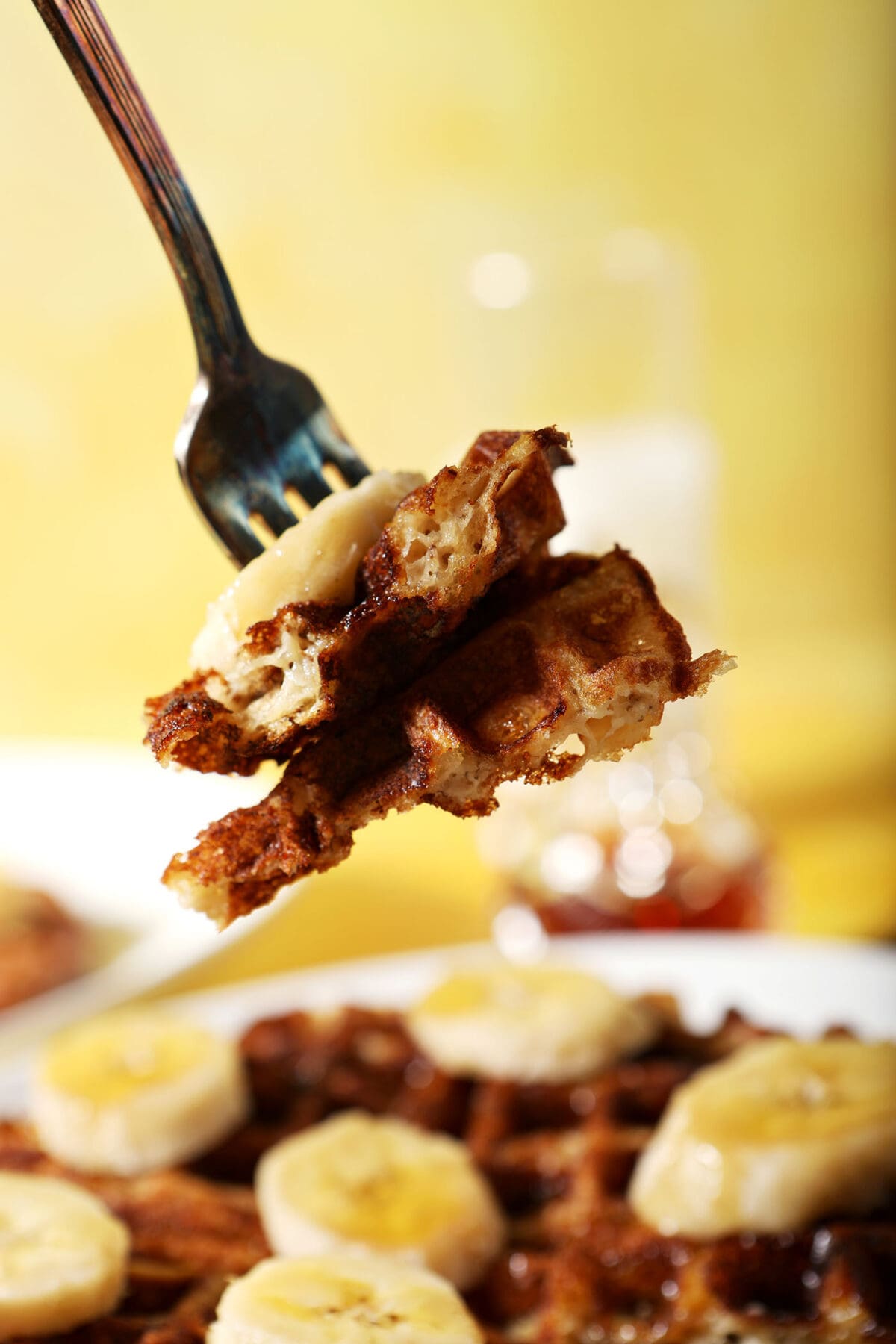 A fork holds a large bite of banana waffles above a plate with a banana waffle on a yellow surface