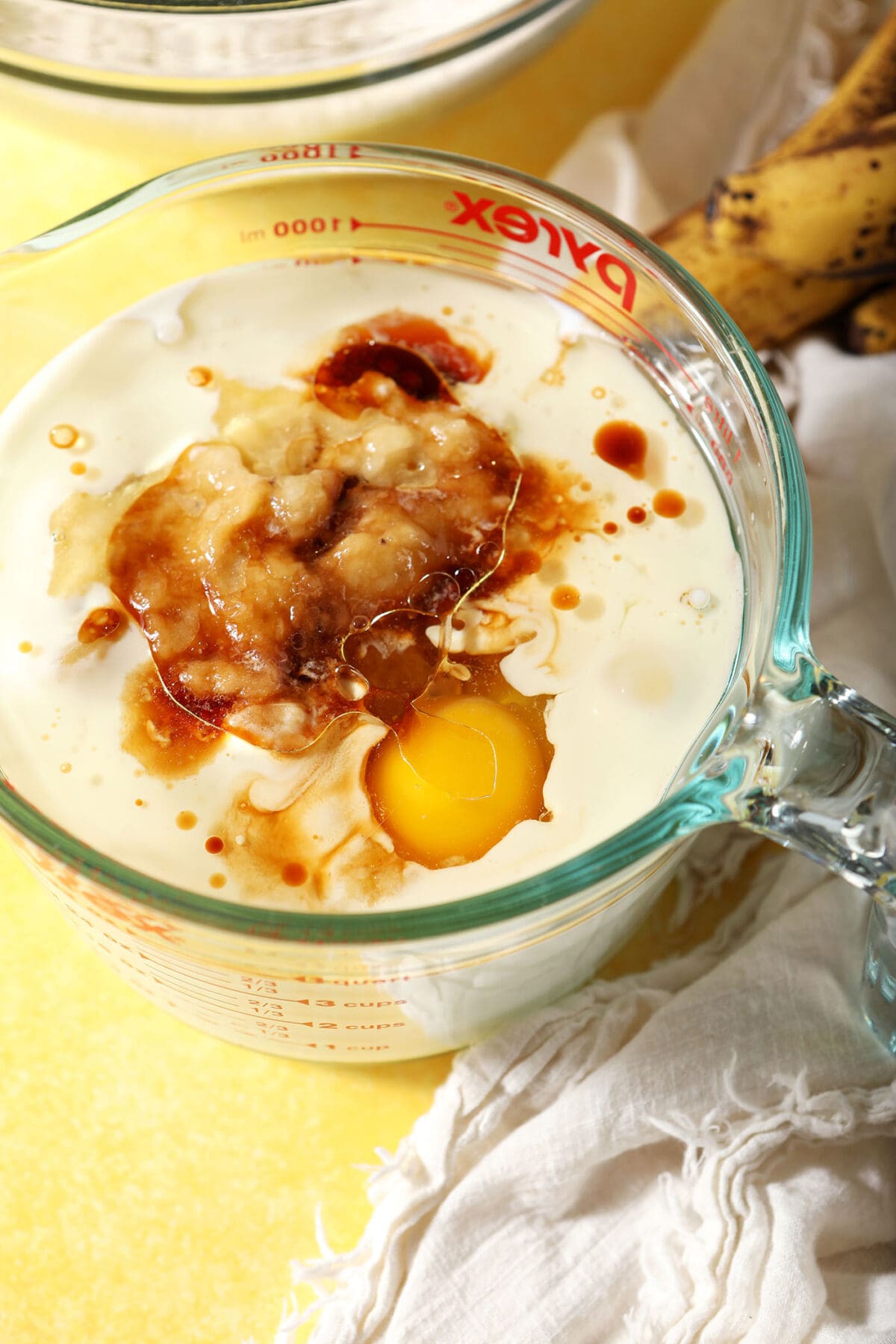 A large measuring cup of wet ingredients on a yellow surface next to a white linen and a few bananas